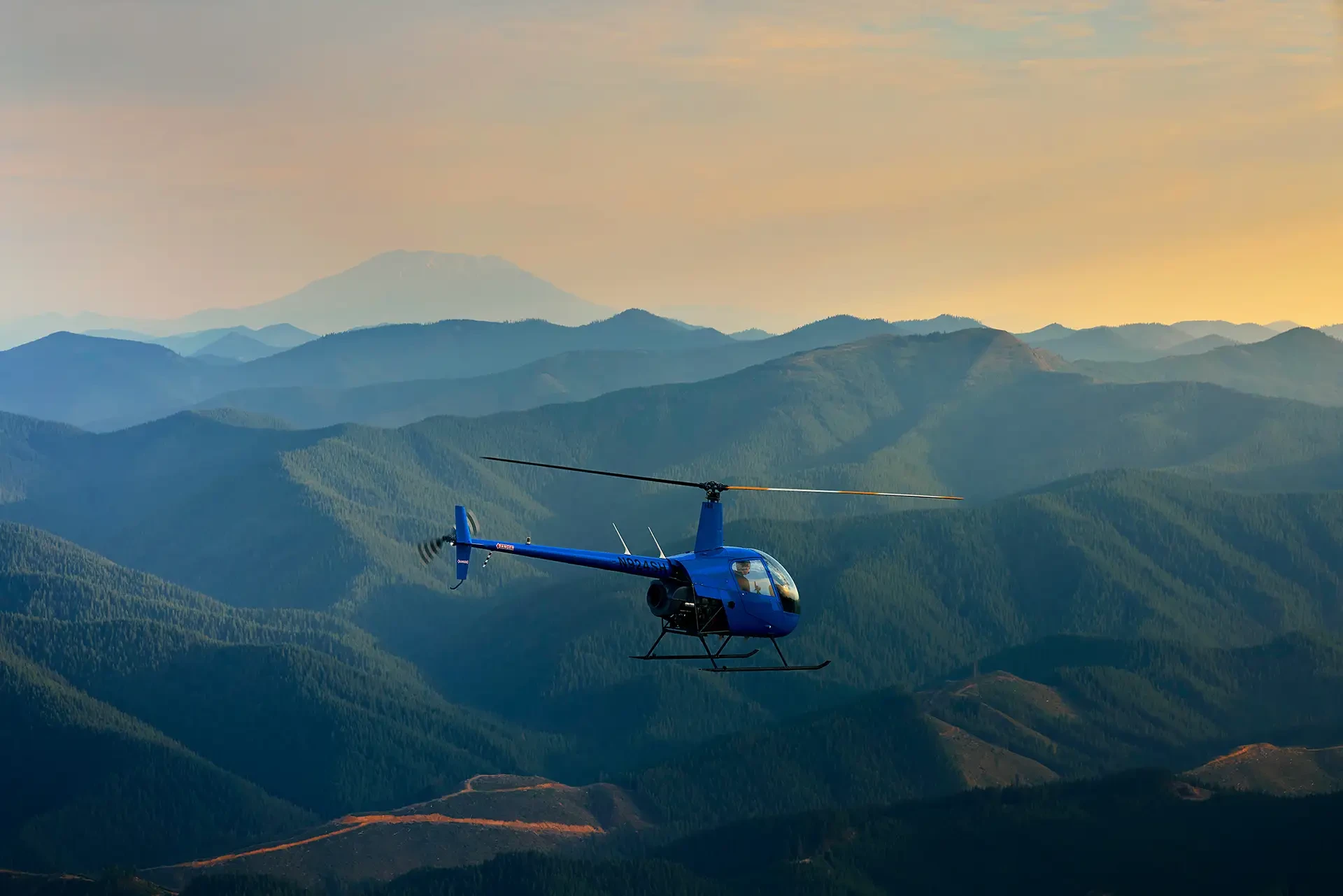 Hillsboro Aero Academy Robinson R22 training helicopter in flight over forested mountain ridges at sunset